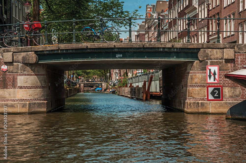 Bridge with iron balustrade and bicycles, old brick buildings and sunny blue sky in Amsterdam. The city is famous for its huge cultural activity, graceful canals and bridges. Northern Netherlands.