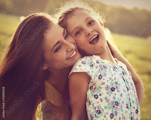 Happy enjoying mother hugging her laughing excited kid girl on sunset bright summer background. Closeup toned emotional portrait.
