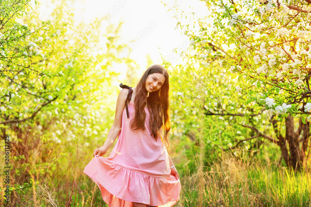 Beatiful smiling girl in pink dress is walking along blooming apple spring garden.