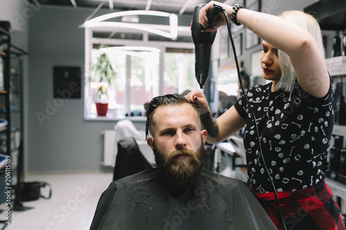 New hairstyle. Side view of young bearded man getting groomed at hairdresser with hair dryer while sitting in chair at barbershop.