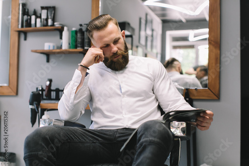 Portrait of handsome bearded man with fashionable hairstyle and beard at barber shop. He chooses a haircut in the journal.