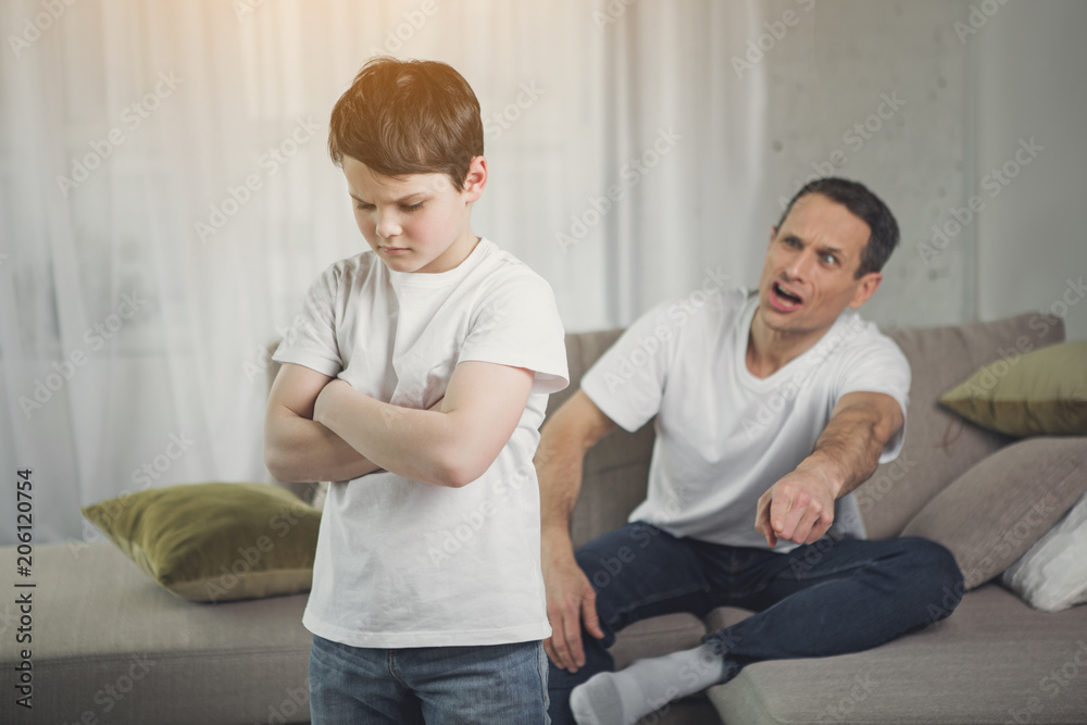 You are punished. Irritated father is yelling at his son. Abused child is standing with crossed arms in room You are punished. Irritated father is yelling at his son. Abused child is standing with crossed arms in room