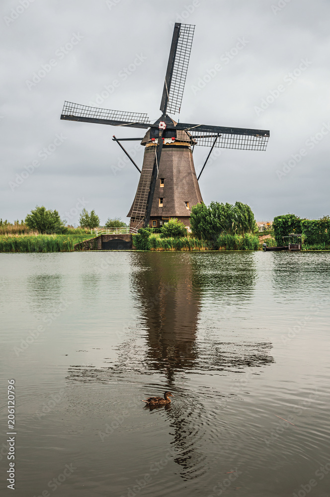 Windmill and bushes reflected on the water of a large canal in a cloudy day at Kinderdijk. Situated in a polder, has the largest concentration of old windmills in the country. Southern Netherlands. Windmill and bushes reflected on the water of a large canal in a cloudy day at Kinderdijk. Situated in a polder, has the largest concentration of old windmills in the country. Southern Netherlands.
