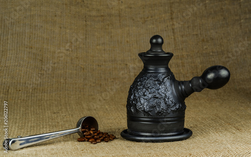 A steel measuring spoon with roasted coffee beans and a made of black clay сoffeepot on a coarse canvas background. photo