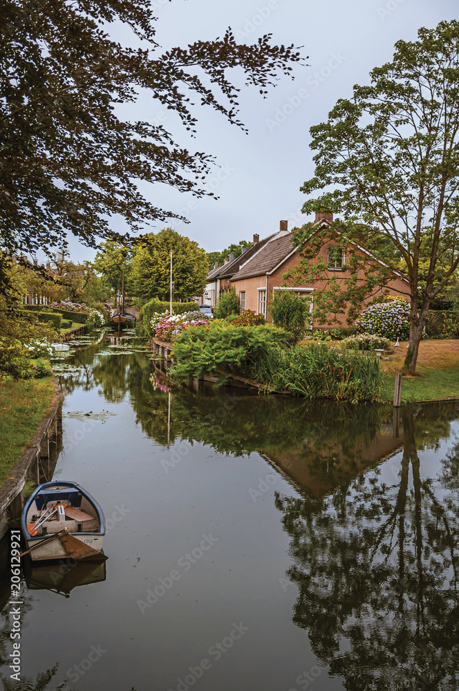 Obraz premium Charming small canal next to rustic houses with lush gardens and trees reflected on the water in cloudy day at Drimmelen. A lovely small hamlet with harbor and elegance streets. Southern Netherlands. 