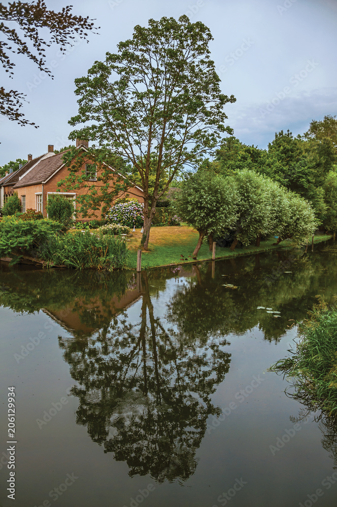 Obraz premium Charming small canal next to rustic house with lush garden and trees reflected on the water in cloudy day at Drimmelen. A lovely small hamlet with harbor and elegance streets. Southern Netherlands. 