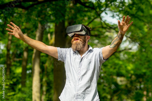 Man with open mouth wearing VR glasses walking in forest, modern technologies concept. Hipster with tattooed arm on natural background. Bearded man exploring surroundings with top-notch gadget