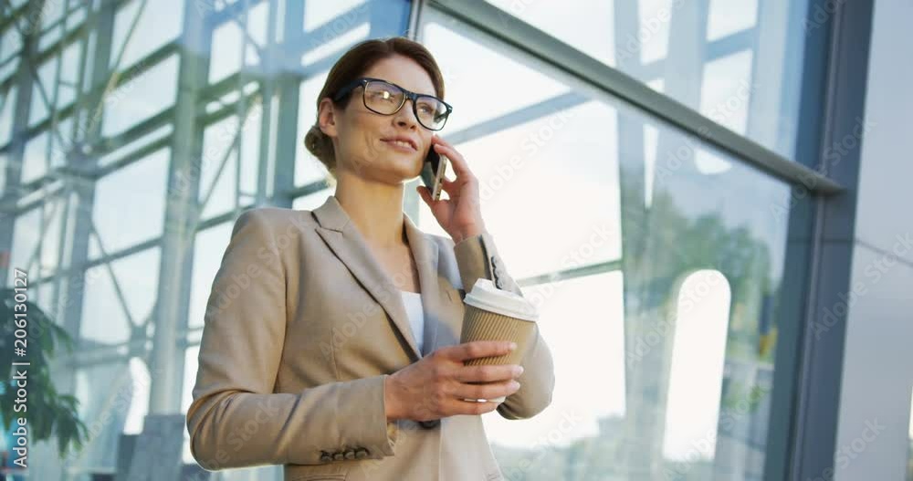 Portrait shot of the attractive smiled businesswoman standing on the glass office center background, talking on the phone and holding her morning coffee on a sunny day. Outdoor Portrait shot of the attractive smiled businesswoman standing on the glass office center background, talking on the phone and holding her morning coffee on a sunny day. Outdoor