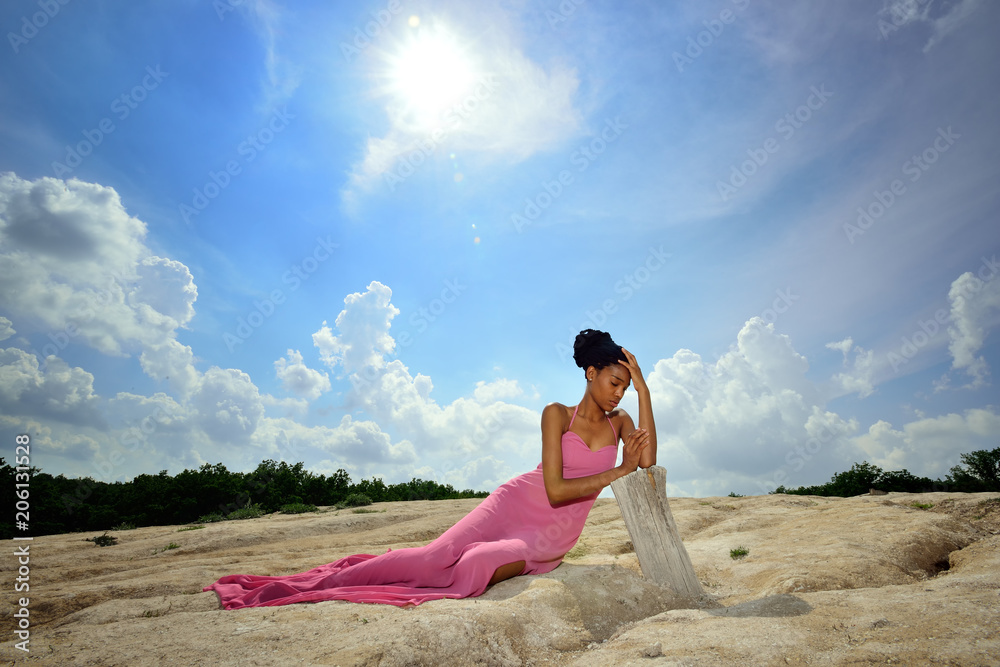 An African American girl is lying in a pink dress on a high hill against the sky, clouds and bright sun and thoughtfully leans her hands on a dry tree. Portrait. Horizontal view