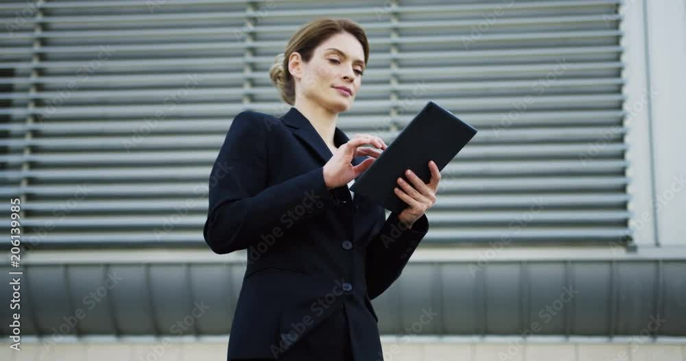 Portrait of the Caucasian good looking successful woman in glasses and black jacket taping on the tablet computer and smiling to the camera near big office building. Outdoors Portrait of the Caucasian good looking successful woman in glasses and black jacket taping on the tablet computer and smiling to the camera near big office building. Outdoors