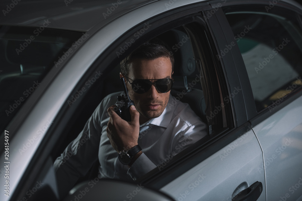 undercover male agent in sunglasses doing surveillance and using talkie walkie in car
