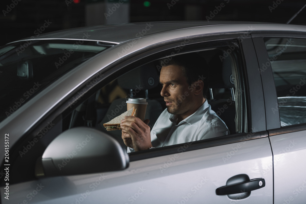 side view of male private detective drinking coffee and eating sandwich in his car