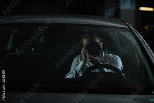 front view of male private detective spying by camera with object glass from his car