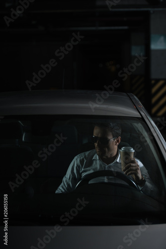 male undercover agent in sunglasses sitting in car with paper cup of coffee