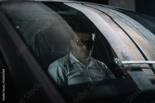 male undercover agent in sunglasses sitting in car with paper cup of coffee while doing surveillance photo