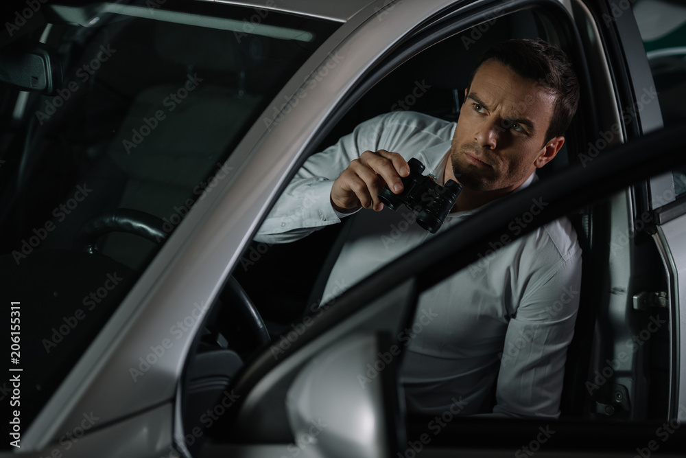 focused undercover male agent doing surveillance by binoculars from his car focused undercover male agent doing surveillance by binoculars from his car