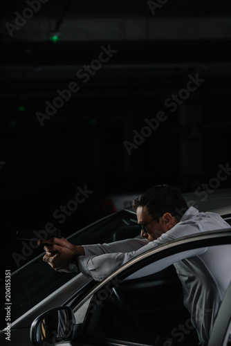 undercover male agent in sunglasses standing near car and aiming by gun