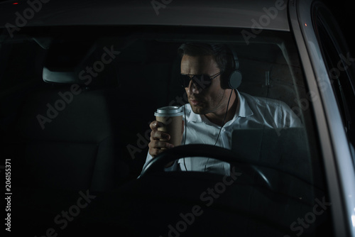 male private detective in headphones doing surveillance with laptop and drinking coffee in car