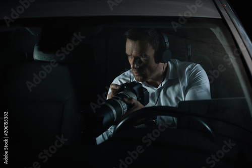 Fotografie concentrated male private detective in headphones spying by camera with lens fro