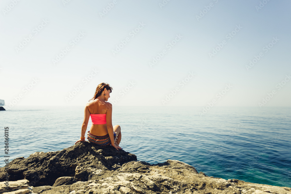 Woman sit on a rock beside the  calm sea. Travel and relax.