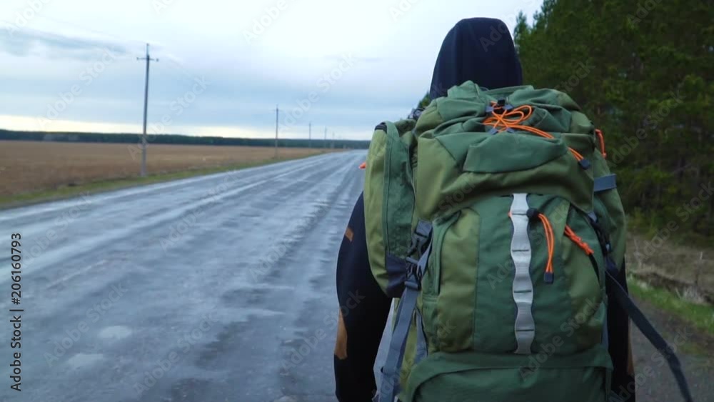 A tourist hitchhiker walks along the road with a backpack on his shoulders. The weather is cloudy and rainy. A tourist hitchhiker walks along the road with a backpack on his shoulders. The weather is cloudy and rainy.
