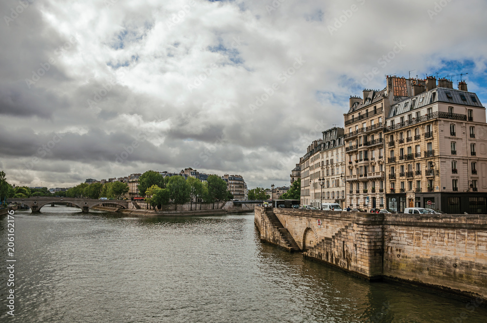 Old buildings, wall on the banks of the Seine River and bridges with cloudy sky in Paris. Known as the “City of Light”, is one of the most impressive world’s cultural center. Northern France.