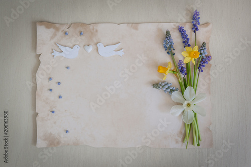 Spring flowers with paper for text and two wooden birds and heart. Wedding, engagement or betrothal concept on on a wooden background.Copy space, top view. Greeting card. Holiday background.