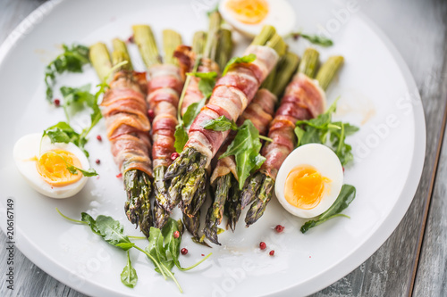 Fresh asparagus wrapped in bacon  on a white plate with arugula tomatoes and eggs