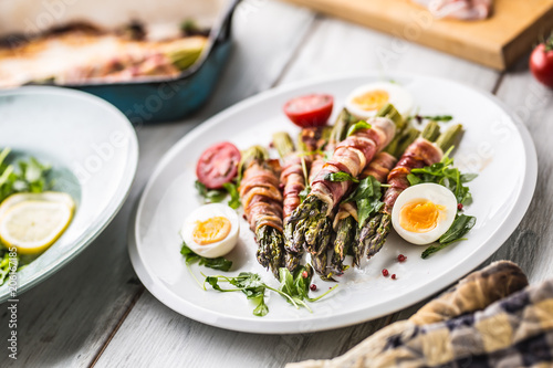 Fresh asparagus wrapped in bacon on a white plate with arugula tomatoes and eggs