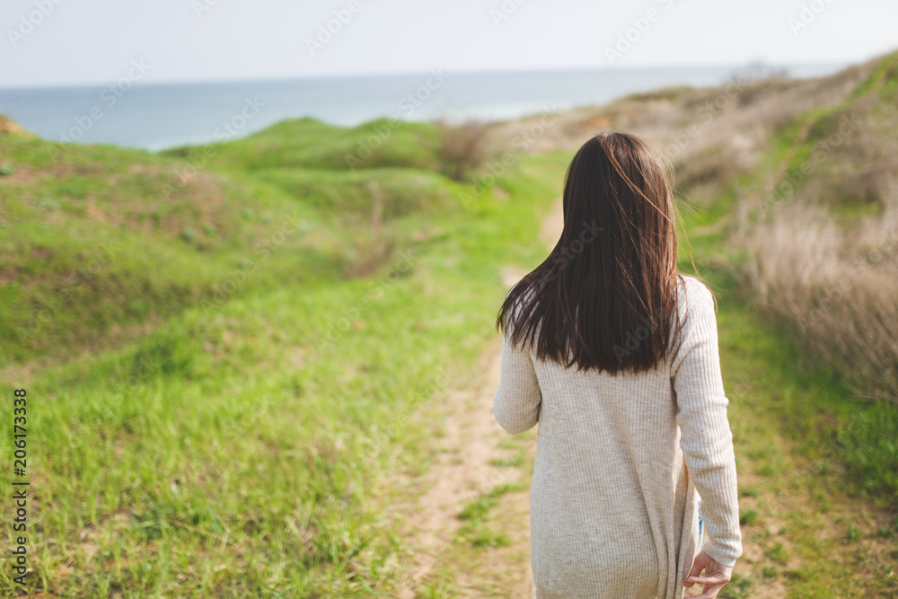 Back view Young brunette woman in light casual clothes walking along path in sunny weather in field near water on green background. Beautiful landscape. Lifestyle, leisure concept. Advertising area. Back view Young brunette woman in light casual clothes walking along path in sunny weather in field near water on green background. Beautiful landscape. Lifestyle, leisure concept. Advertising area.