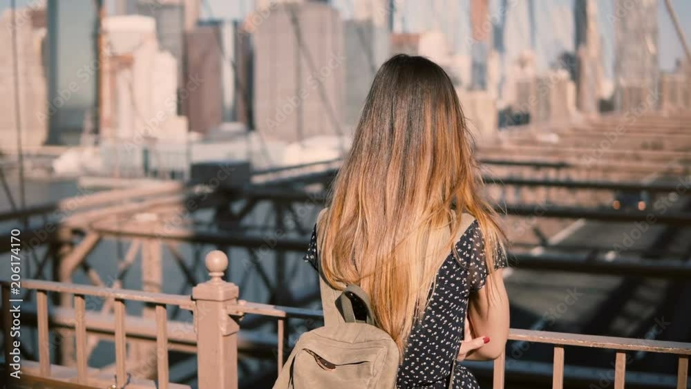 Young female Caucasian tourist in stylish sunglasses with backpack standing alone at Brooklyn Bridge, New York City 4K.
