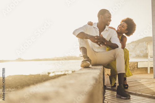 black race african couple in love and vacation sit down enjoying together with big smiles and laugh. casual clothes like fashion style with nice sunset backlight on the background. tenerife location. photo