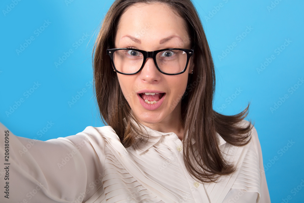 Fototapeta premium Young woman in glasses makes a selfie portrait on a blue background.