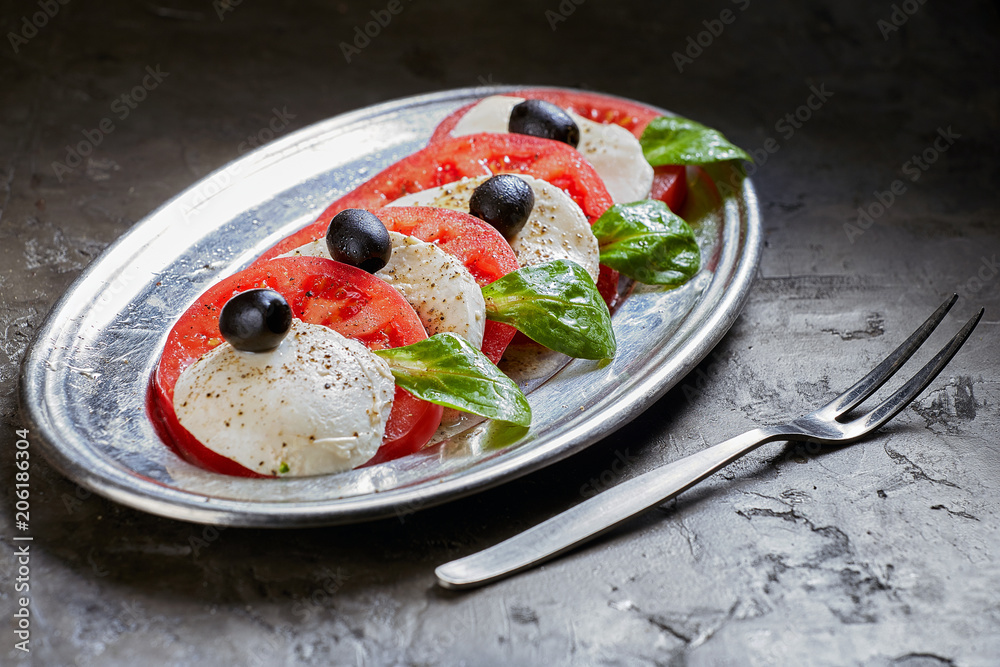 Caprese Salad. Tomato and Mozzarella slices, Basil and olives is on a plate. Dark background.