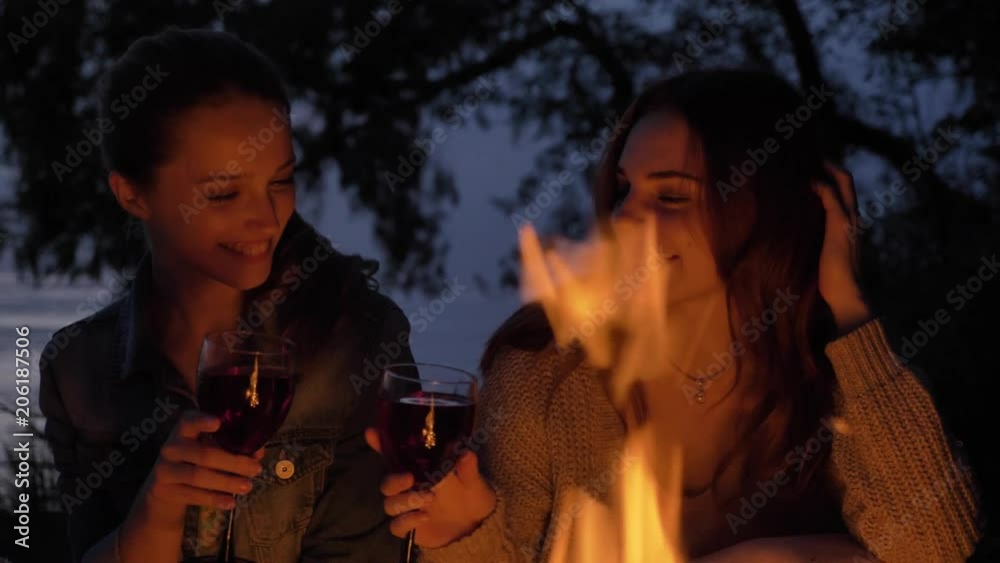 Two young Caucasian girls sit in the evening by the fire in nature, drink wine, smiling Two young Caucasian girls sit in the evening by the fire in nature, drink wine, smiling