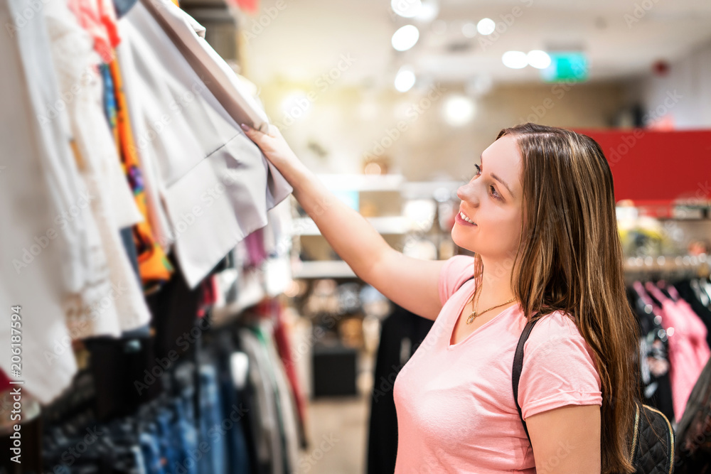 Young woman shopping in clothing store. Happy girl looking at clothes in  shop at mall. Smiling student choosing outfit. Stock Photo