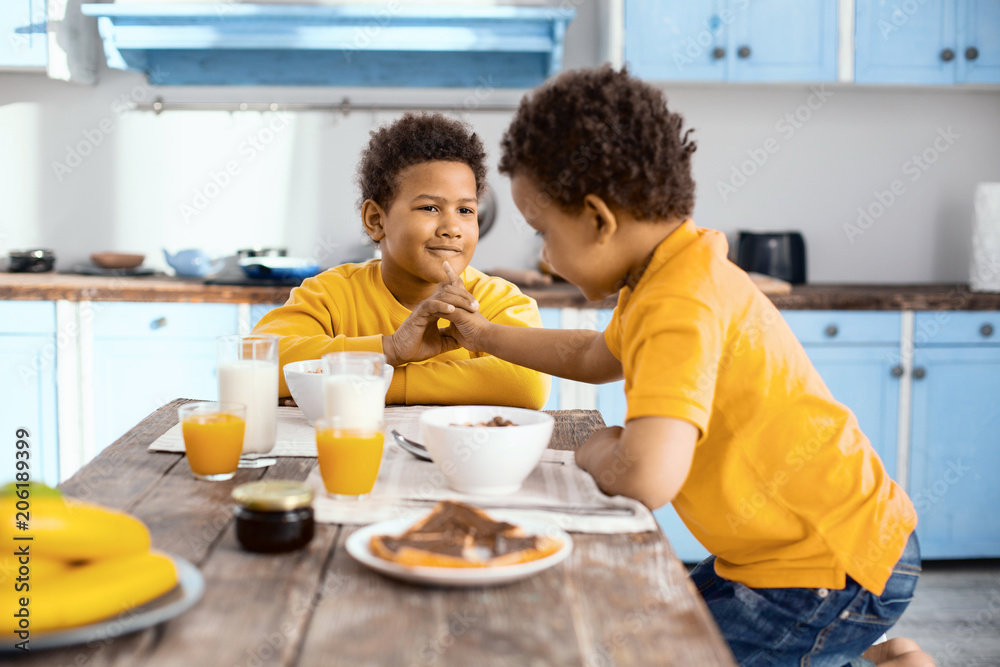 Fun at breakfast. Curly-haired little boy sitting at the table next to his elder brother and teasing him, trying to poke his chin while they having breakfast
