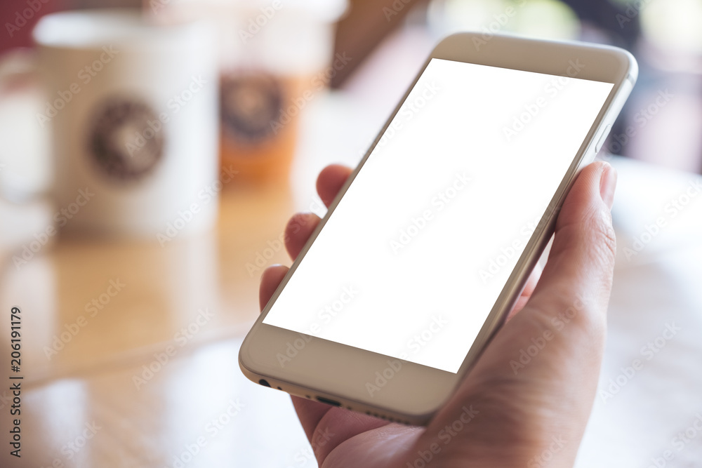 Mockup image of hand holding white mobile phone with blank desktop screen on table in cafe