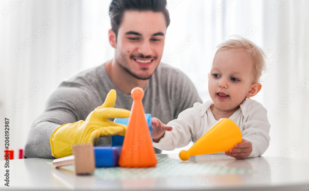Handsome man in yellow gloves cleaning table while his little child playing toys, modern father Handsome man in yellow gloves cleaning table while his little child playing toys, modern father