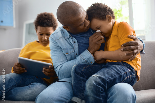 Daily care. Caring young father sitting on the sofa, hugging his crying little son tightly and wiping his nose while the older boy playing on tablet