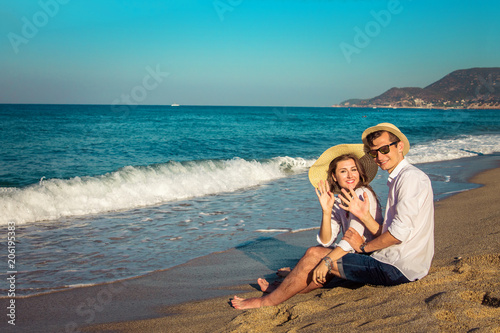 Young happy couple sitting on tropical beach and waving at camera. Travellers chilling and relaxing
