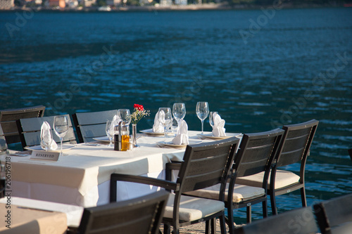Romantic dinner by sea restaurant with beautiful view on mountains. Served table with white and beige tableclothes, wineglasses of ice champagne, red flowers.