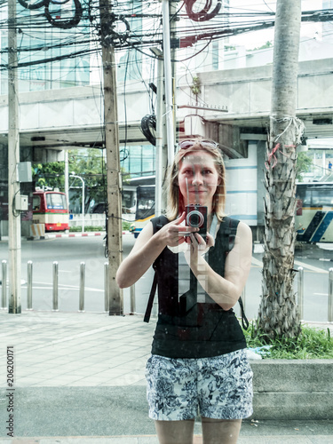 Blonde girl takes photo in the mirroring glass window in a city
