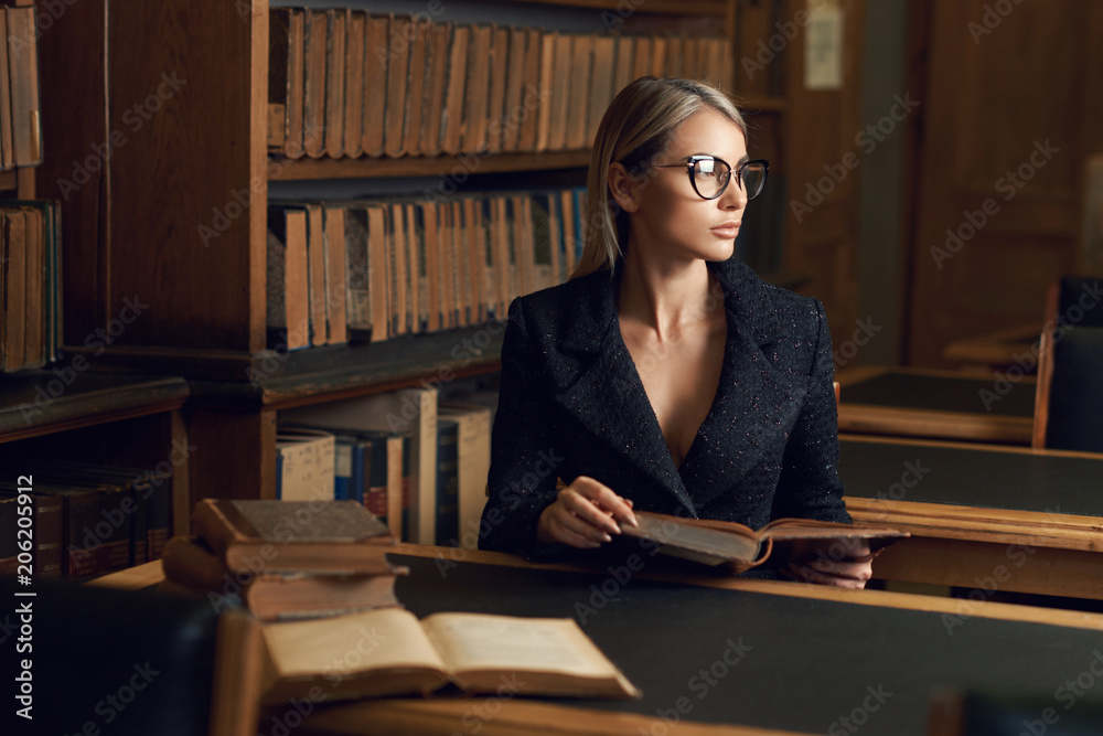 Beautiful blonde woman wearing elegant black tweed jacket and glasses  sitting at desk beside bookshelf and reading book. Young gorgeous female  student studying at library. Smart is new sexy concept. Stock-Foto