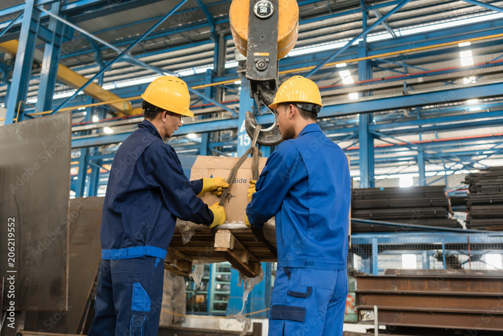 Two Asian workers handling heavy loading lifted by crane in the interior of a metallurgical factory Two Asian workers handling heavy loading lifted by crane in the interior of a metallurgical factory