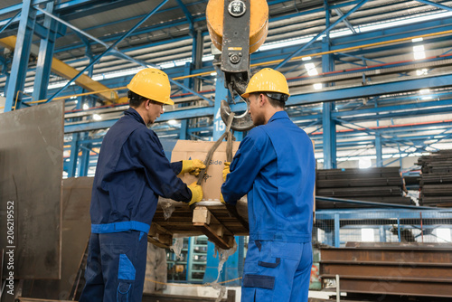 Two Asian workers handling heavy loading lifted by crane in the interior of a metallurgical factory