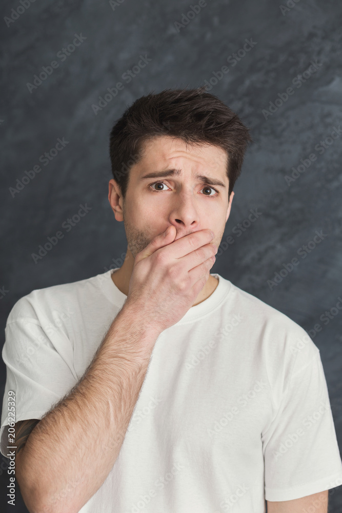 Frightened man covering mouth with hand at gray background