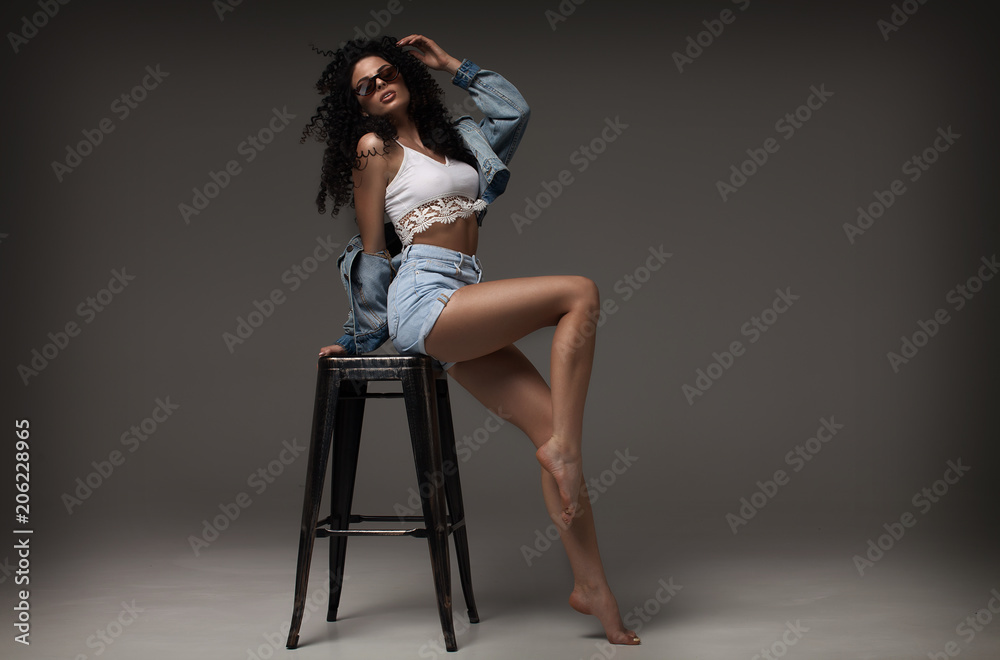 Sexy attractive brunette woman posing in fashionable jeans in studio. Fashion model posing. Long curly hair. Sexy attractive brunette woman posing in fashionable jeans in studio. Fashion model posing. Long curly hair.