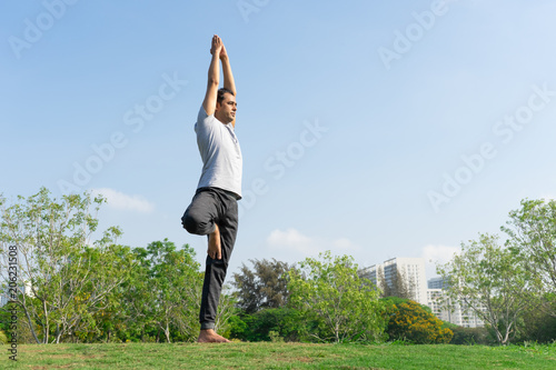 Indian male yoga instructor standing in tree pose on green lawn photo