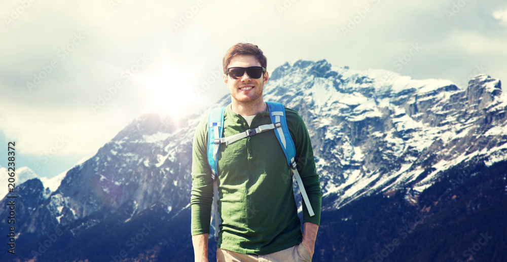 travel, tourism and people concept - happy young man in sunglasses with backpack over alps mountains background